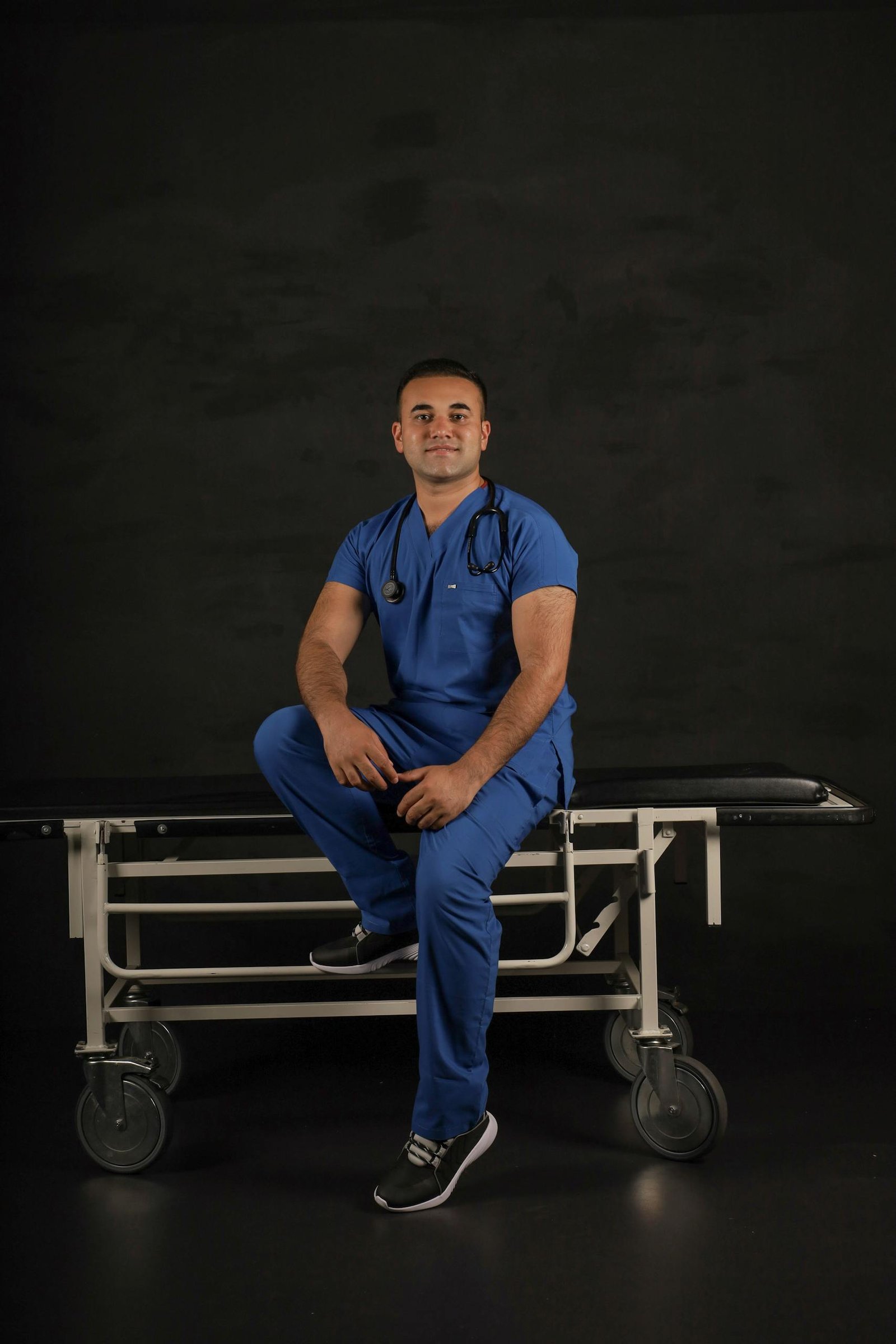 Male doctor in blue scrub suit seated on a stretcher with a stethoscope, smiling on a dark studio background.